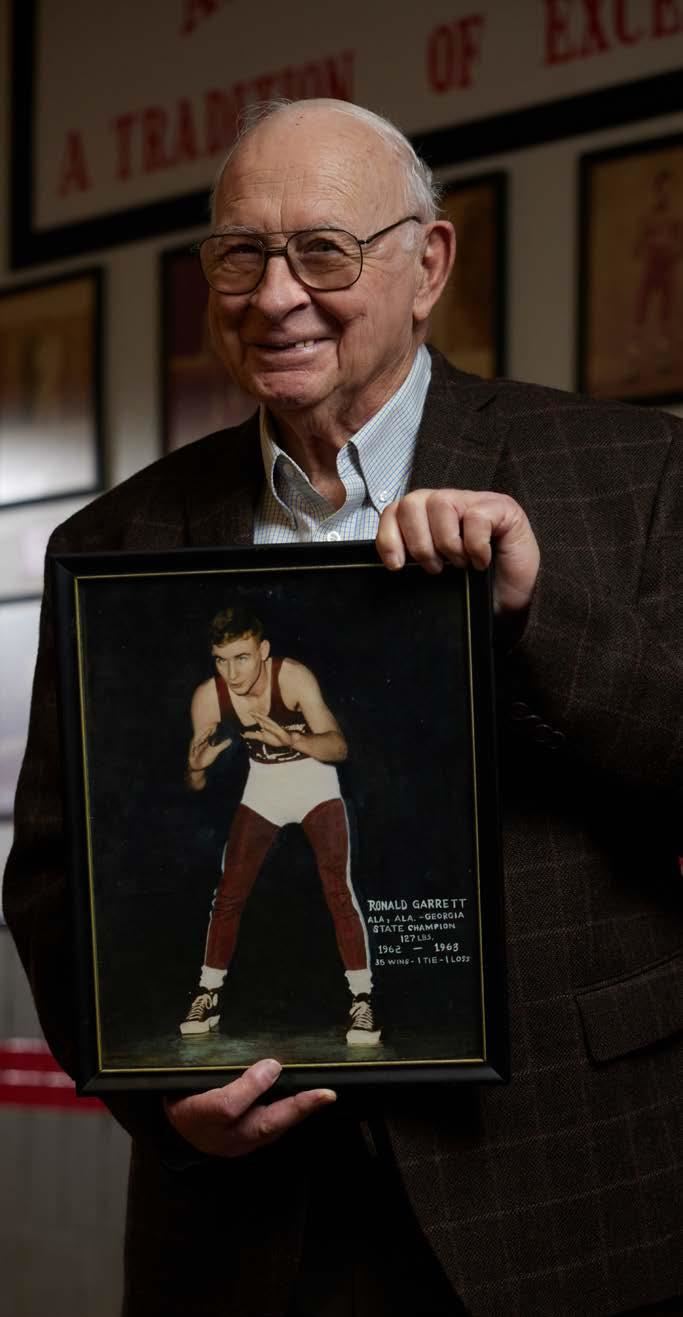  Ronald Garrett holding a framed photo of himself as a young wrestler in a maroon and white uniform.